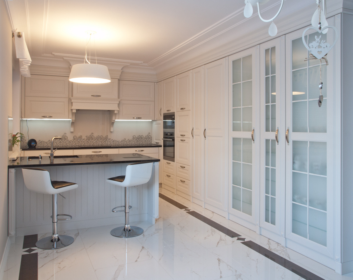 Elegancka, jasna kuchnia z wyspą i dwoma białymi hokerami, białe szafki z przeszkleniami, marmurowa podłoga z czarnymi akcentami i oświetlenie LED pod szafkami.