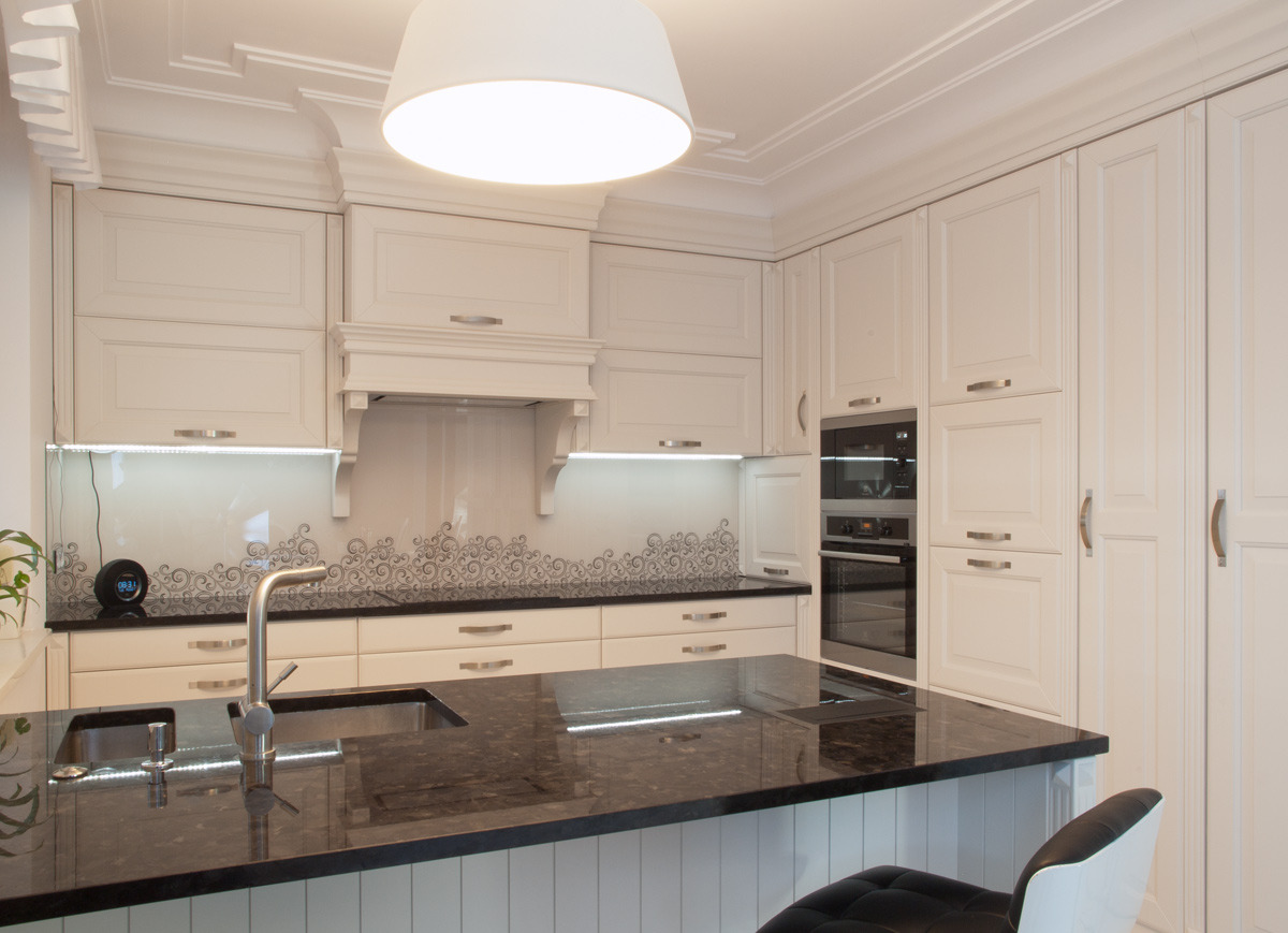 Elegancka kuchnia w odcieniach bieli z ciemnym, błyszczącym blatem i dekoracyjnym panelem nad blatem, oświetlona lampą wiszącą i listwami LED pod szafkami.