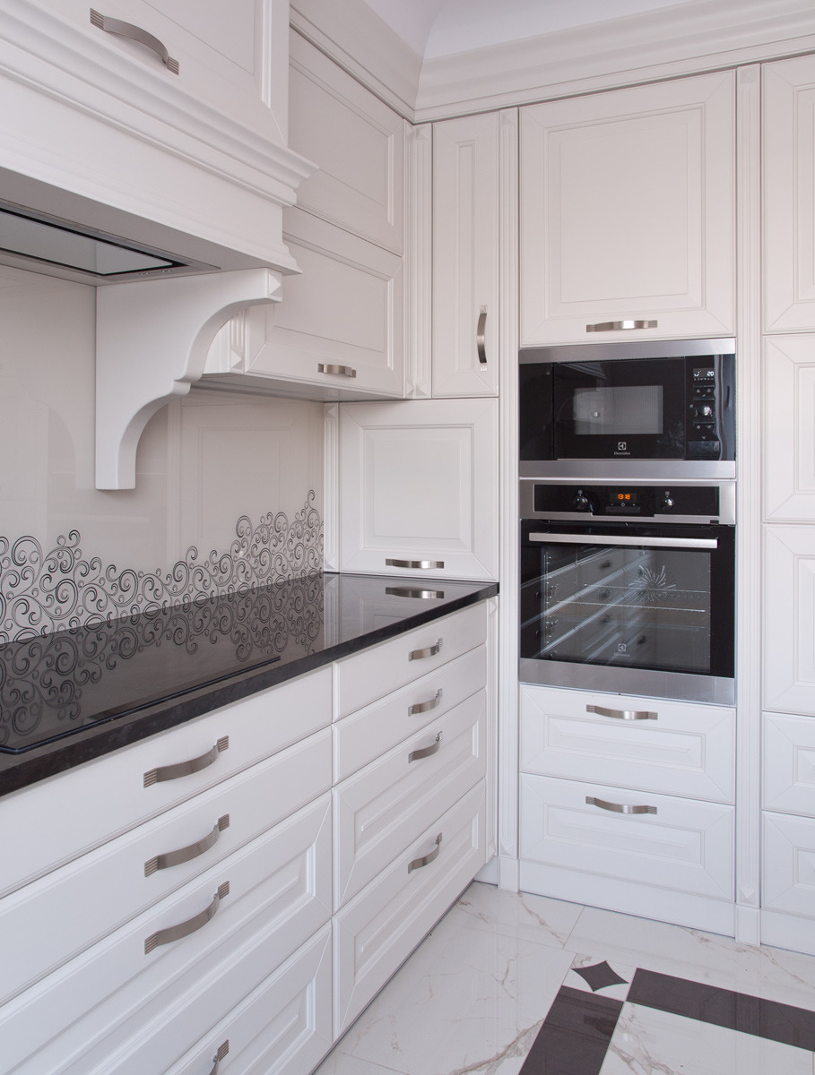 Klasyczna biała zabudowa kuchenna z czarnym blatem i sprzętem AGD marki Electrolux, widok z perspektywy narożnika, wzorzysta mozaika na ścianie nad blatem.