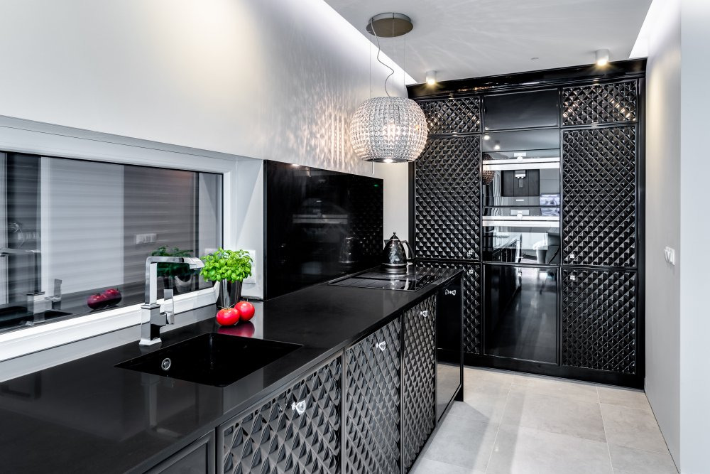 Nowoczesna, czarna kuchnia z błyszczącymi frontami szafek o geometrycznym wzorze, okrągła lampa wisząca z kryształkami, czarny blat, zlew i bateria, bazylia w doniczce i dwa czerwone pomidory...