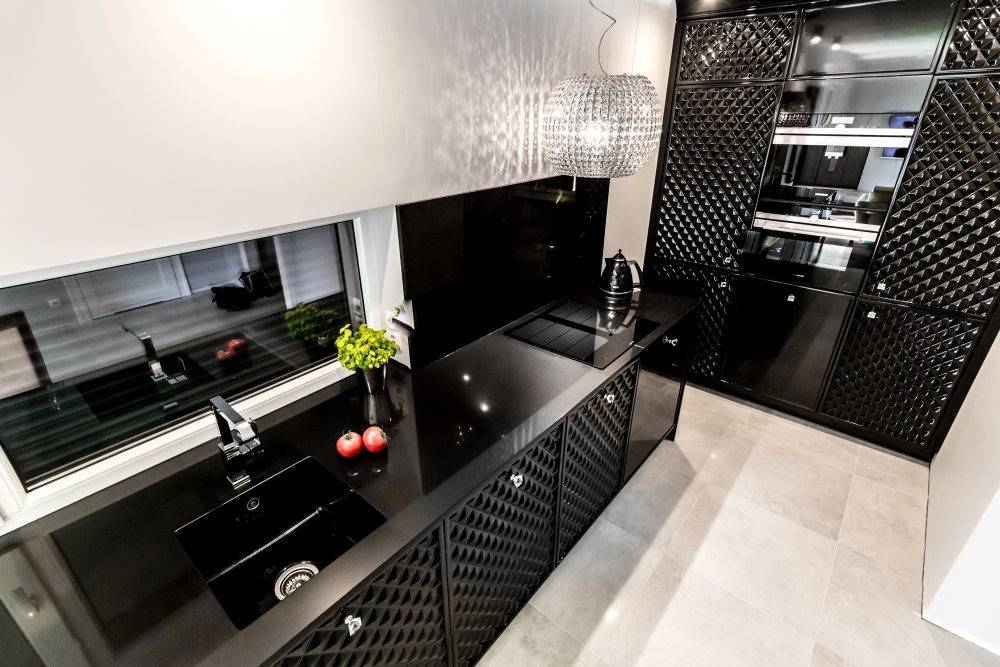 Elegancka, czarna kuchnia z błyszczącymi frontami szafek o fakturze rombów, czarnym blatem, nowoczesną lampą wiszącą i widokiem z okna na zieleń.