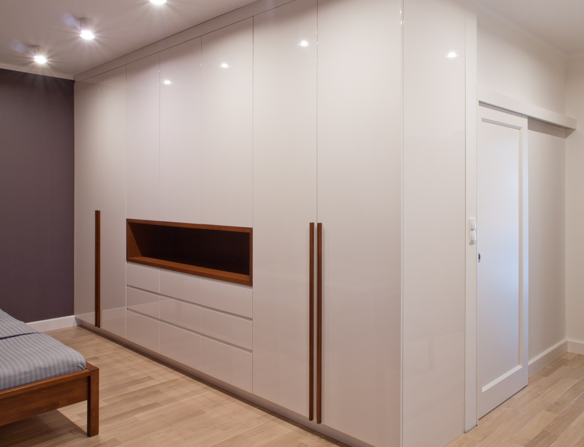 Biała, lakierowana szafa wnękowa z drewnianymi uchwytami i otwartą półką w kolorze drewna, widoczna w sypialni z łóżkiem i przesuwnymi drzwiami.