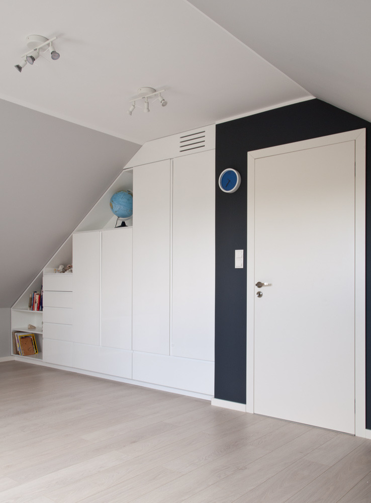 Biała szafa na wymiar zabudowana pod skosem dachu, z otwartymi półkami, globusem i dekoracjami, obok ściana pomalowana na granatowo z białymi drzwiami, oświetlenie punktowe na suficie.