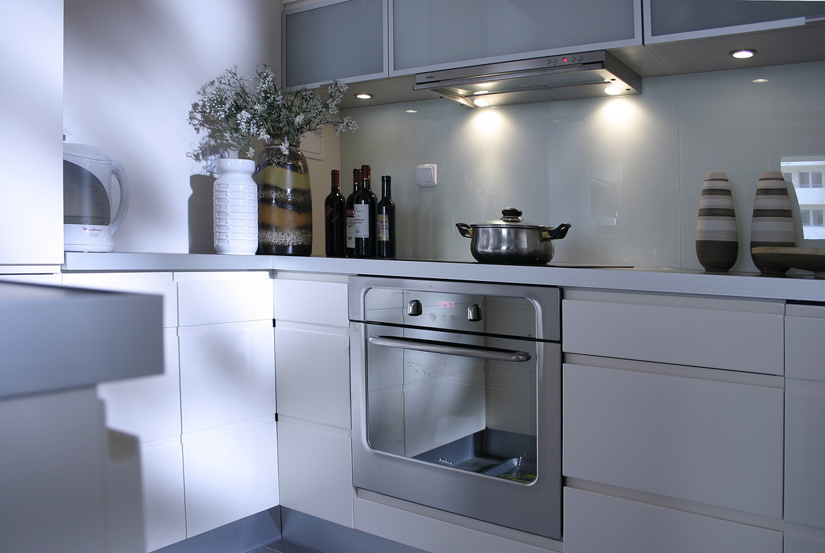Nowoczesna, minimalistyczna kuchnia z białymi frontami szafek, srebrnym piekarnikiem, szklanym panelem nad blatem, dekoracyjnymi wazonami i butelkami wina, oświetlona punktowymi lampami.