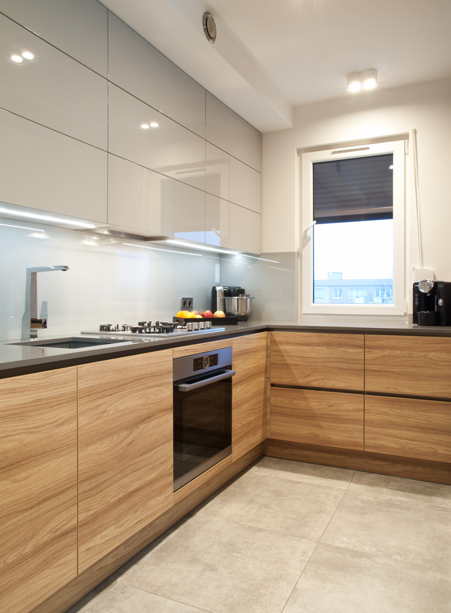 Nowoczesna kuchnia w kształcie litery L z drewnianymi frontami szafek dolnych i białymi, lakierowanymi szafkami górnymi, szary blat i płytki podłogowe, oświetlenie LED pod szafkami.