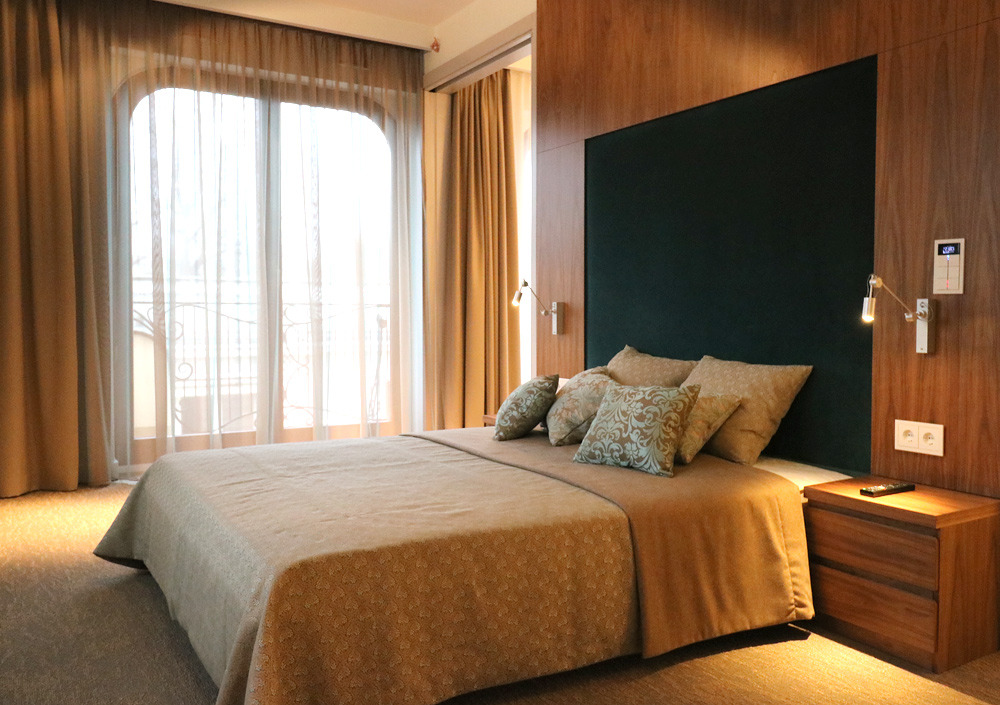 Nowoczesna sypialnia z łóżkiem małżeńskim, ciemnozielonym zagłówkiem, drewnianą boazerią, zasłonami w oknach balkonowych i stolikiem nocnym z pilotem.