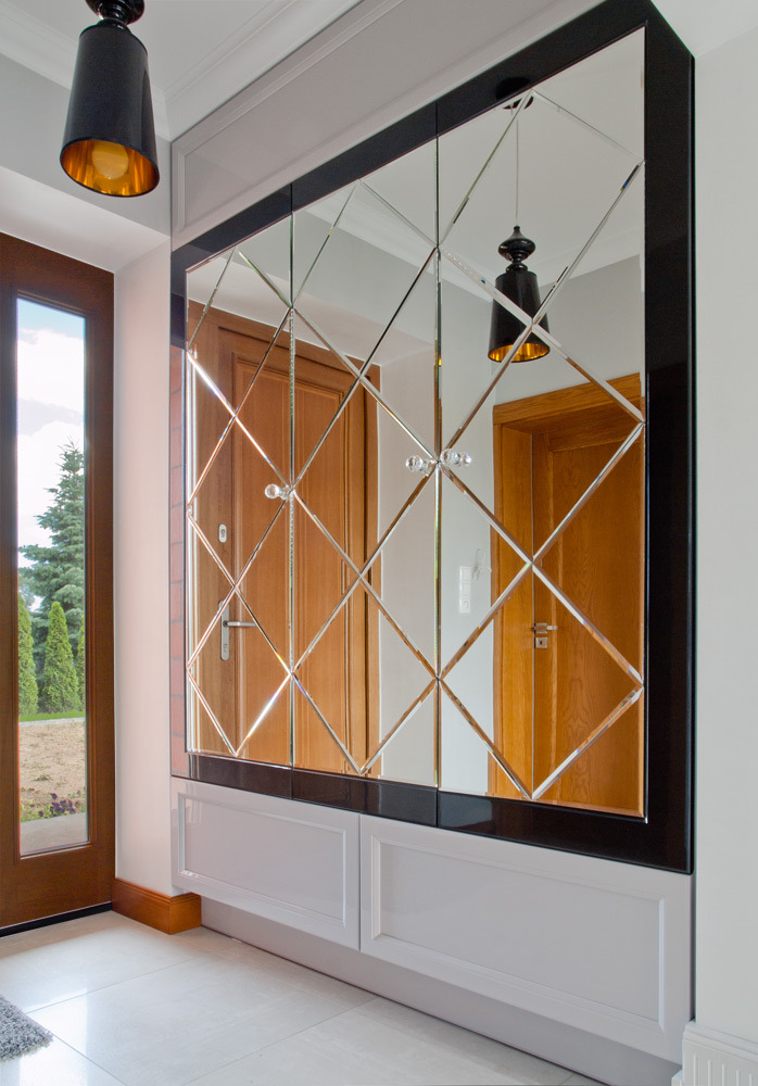 Lustrzana ściana z dekoracyjnymi szprosami w przedpokoju, odbijająca drzwi i lampę wiszącą, z dolną zabudową w kolorze białym, podłoga z jasnych płytek.