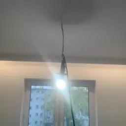 Przygotowanie do malowania sufitu: żarówka na kablu oświetla wnętrze, fragment ściany z widocznymi śladami po wcześniejszych pracach, okno z widokiem na budynek mieszkalny.