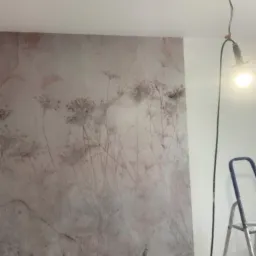 Ściana z tapetą w kwiatowy wzór w odcieniach beżu i szarości, oświetlona lampą roboczą, z drabinką malarską w tle.