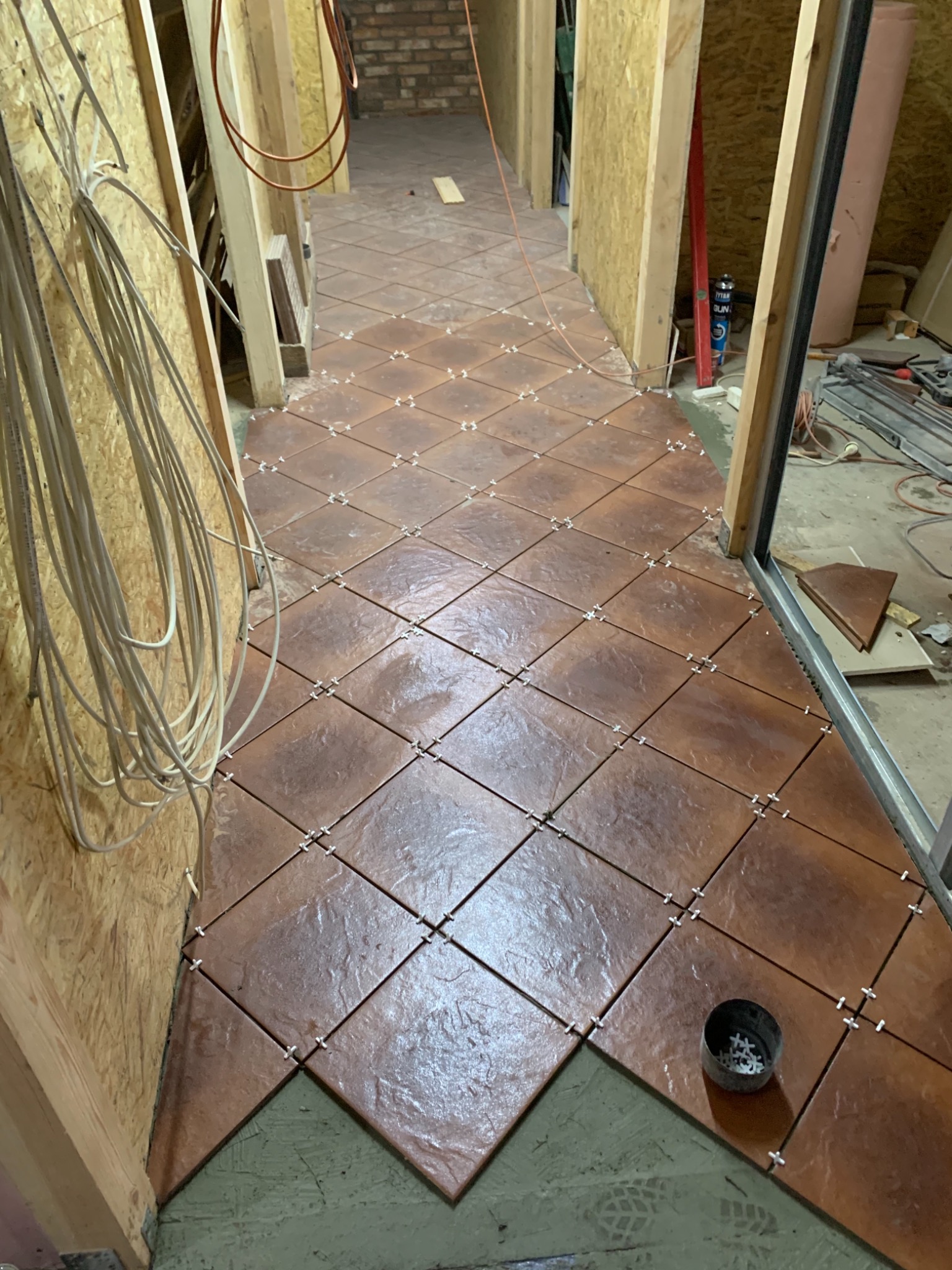 Układanie brązowych płytek podłogowych w romby z użyciem krzyżyków dystansowych w korytarzu w Myśliborzu, widoczne przewody elektryczne i narzędzia pracy.