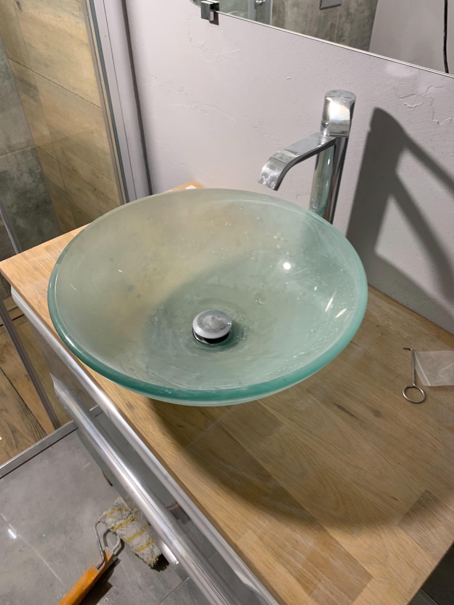 Szklana umywalka nablatowa w odcieniach zieleni z chromowaną baterią, zamontowana na drewnianym blacie. W tle widoczna kabina prysznicowa i fragment pomalowanej ściany. Na blacie leżą akcesoria...