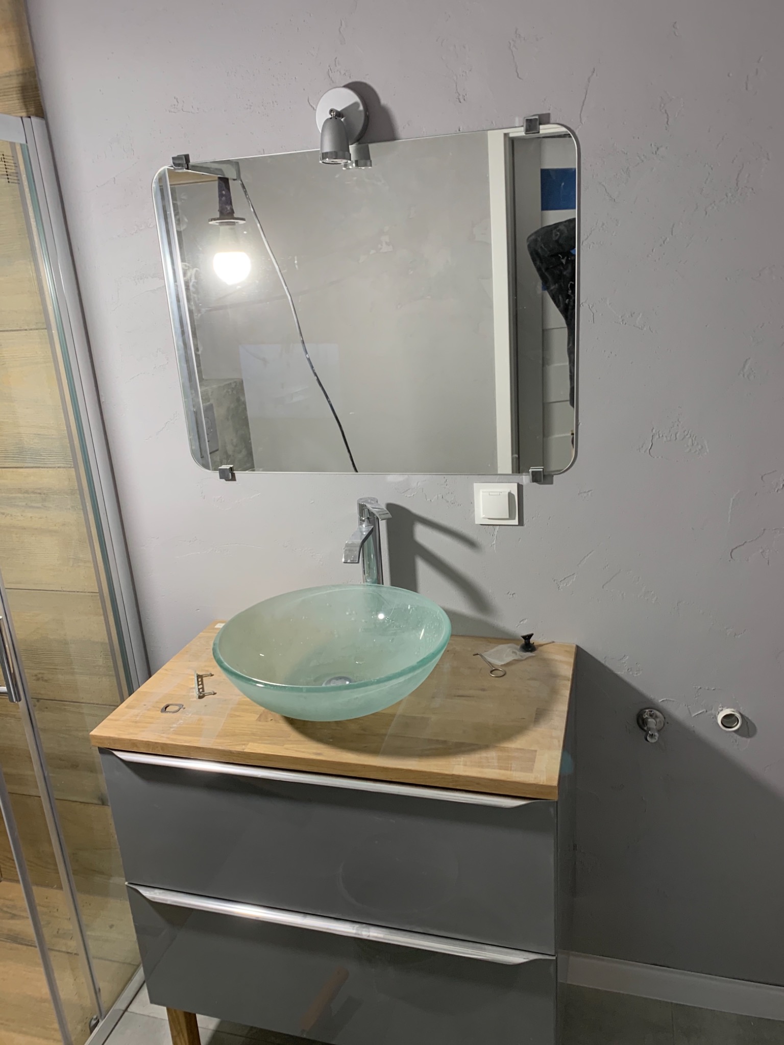 Szklana umywalka w łazience z szarą ścianą o fakturze baranka, lustro z oświetleniem, widoczne narzędzia na blacie, kabina prysznicowa z drewnopodobnymi płytkami w tle.