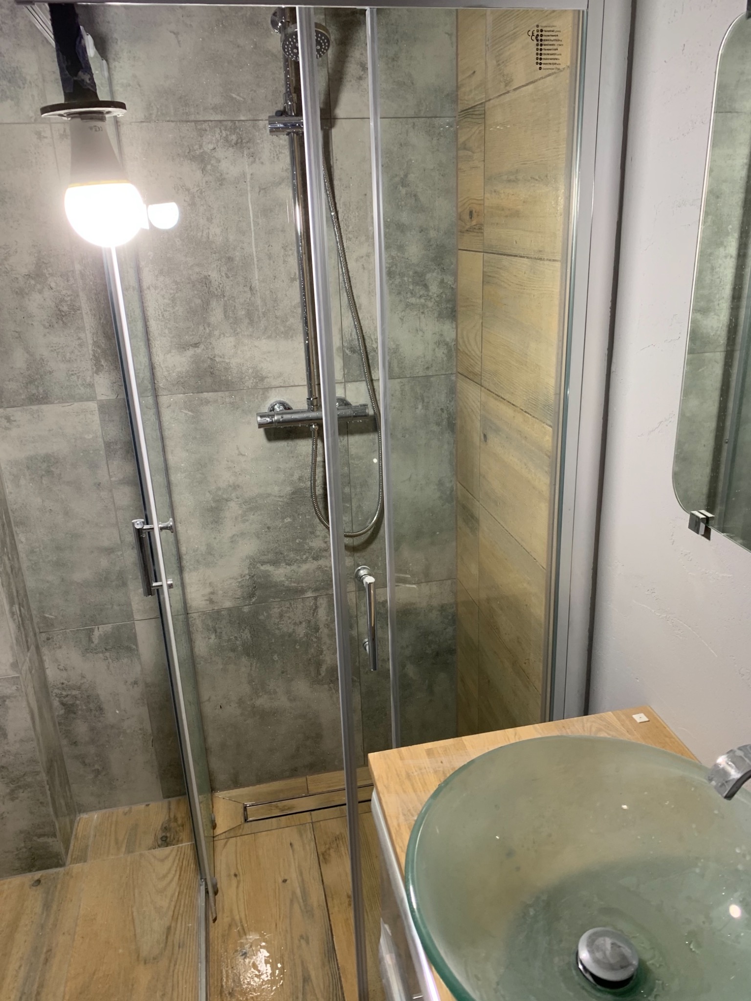 Szklana kabina prysznicowa z szarymi płytkami imitującymi beton, drewnopodobne płytki na podłodze, przezroczysta umywalka na drewnianym blacie, widoczny fragment baterii.