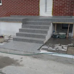 Granitowe schody wejściowe do domu z cegły, zabezpieczone drzwi wejściowe, widoczne narzędzia i wiaderka z zaprawą, trwają prace wykończeniowe.