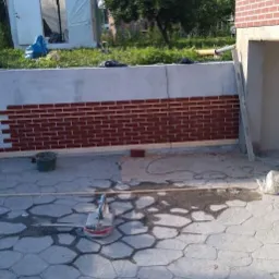 Częściowo wyłożona cegłą elewacja budynku z widocznym narzędziem do cięcia płytek na pierwszym planie i szarą kostką brukową na podłożu.