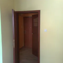 Wnętrze mieszkania z widocznym, częściowo otwartym, brązowym skrzydłem drzwiowym z szybką, prowadzącym do drugiego pomieszczenia z zamkniętymi drzwiami. Ściany w kolorze kremowym i jasnozielonym.