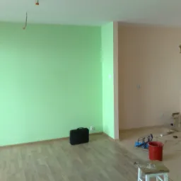 Świeżo pomalowane na jasnozielono ściany pokoju z drewnianą podłogą, widoczny czarny neseser przy ścianie i narzędzia malarskie w sąsiednim pomieszczeniu z beżowymi ścianami.