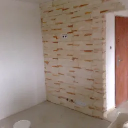 Ściana wykończona dekoracyjną okładziną imitującą kamień, z widocznymi gniazdkami elektrycznymi i puszkami instalacyjnymi, częściowo pomalowana ściana obok i wiadro z farbą na podłodze.