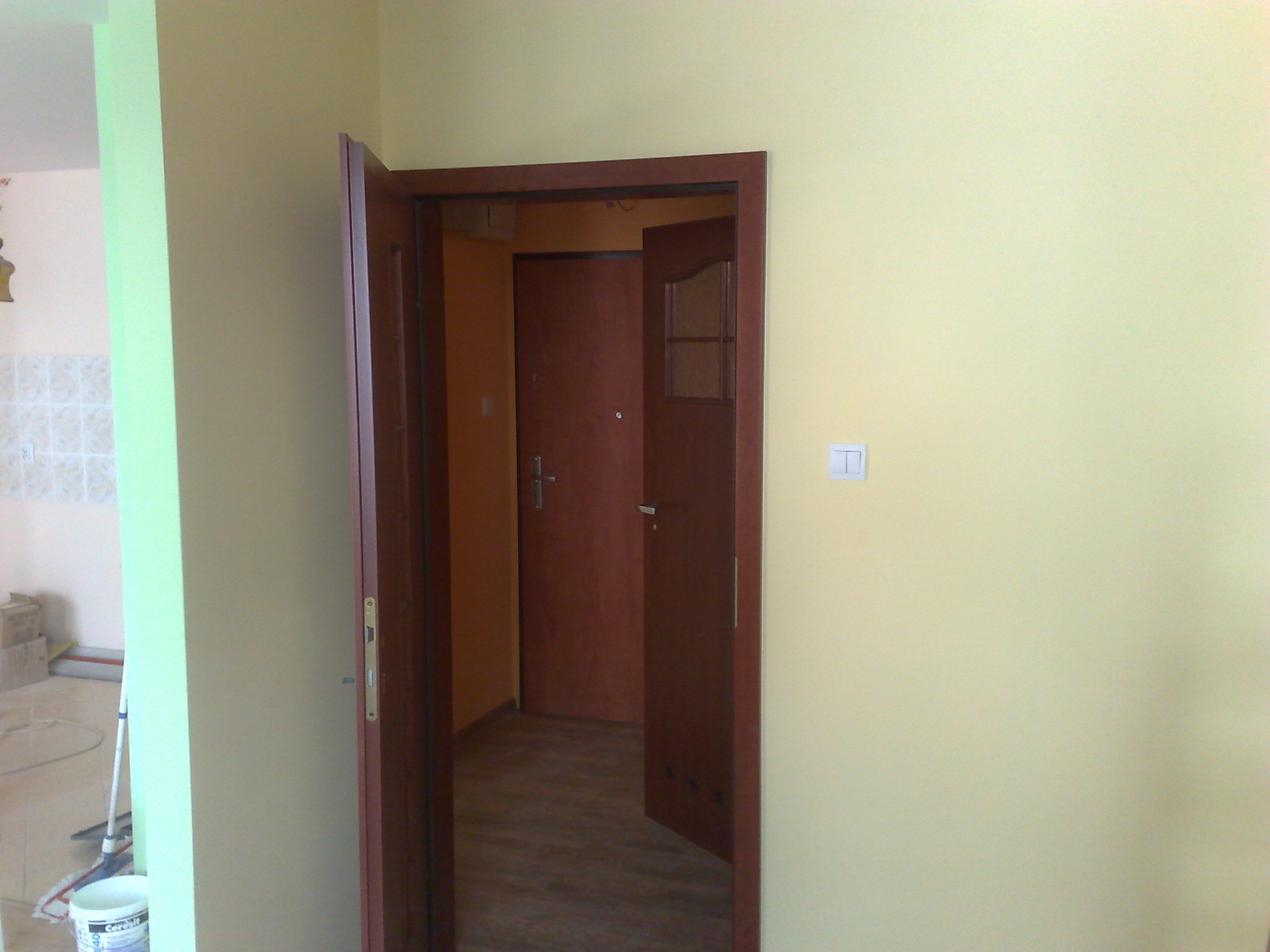 Wnętrze mieszkania z widocznym, częściowo otwartym, brązowym skrzydłem drzwiowym z szybką, prowadzącym do drugiego pomieszczenia z zamkniętymi drzwiami. Ściany w kolorze kremowym i jasnozielonym.