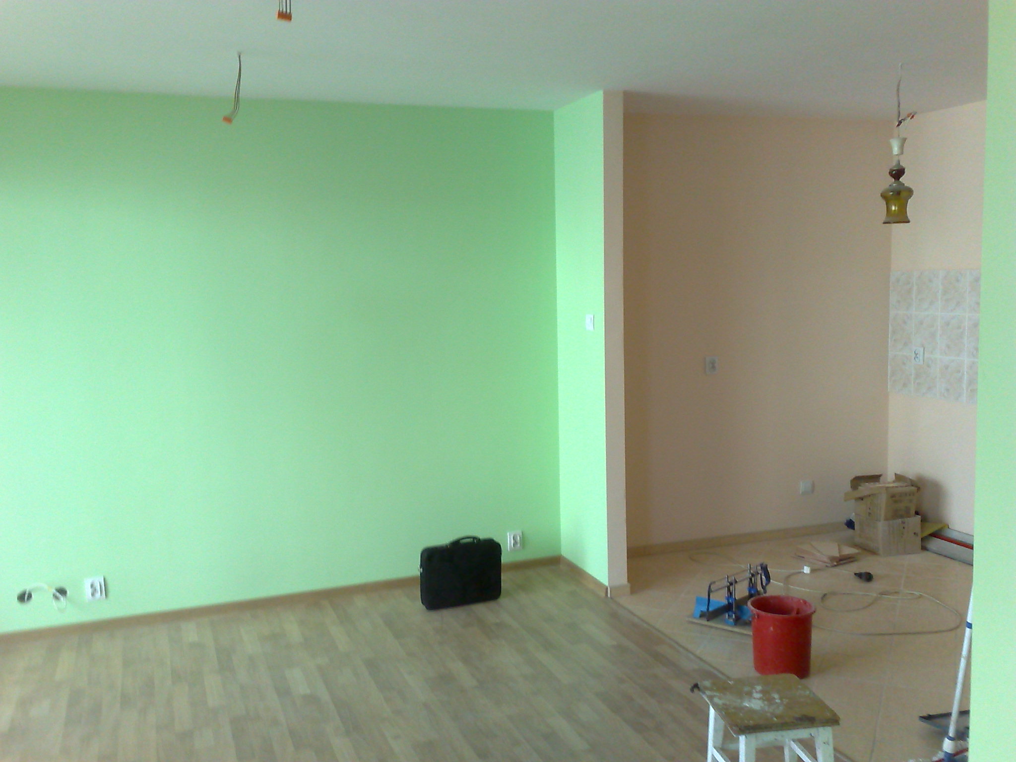 Świeżo pomalowane na jasnozielono ściany pokoju z drewnianą podłogą, widoczny czarny neseser przy ścianie i narzędzia malarskie w sąsiednim pomieszczeniu z beżowymi ścianami.