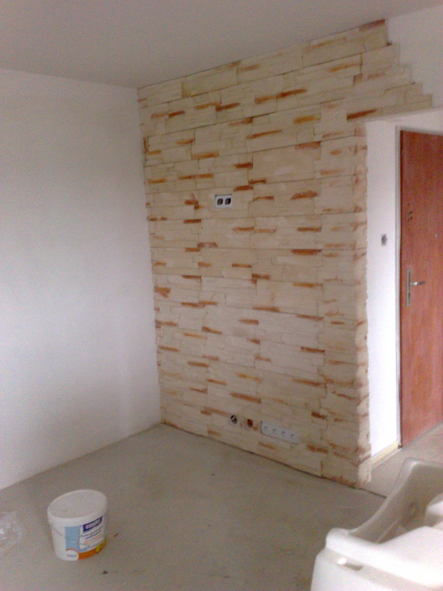 Ściana wykończona dekoracyjną okładziną imitującą kamień, z widocznymi gniazdkami elektrycznymi i puszkami instalacyjnymi, częściowo pomalowana ściana obok i wiadro z farbą na podłodze.