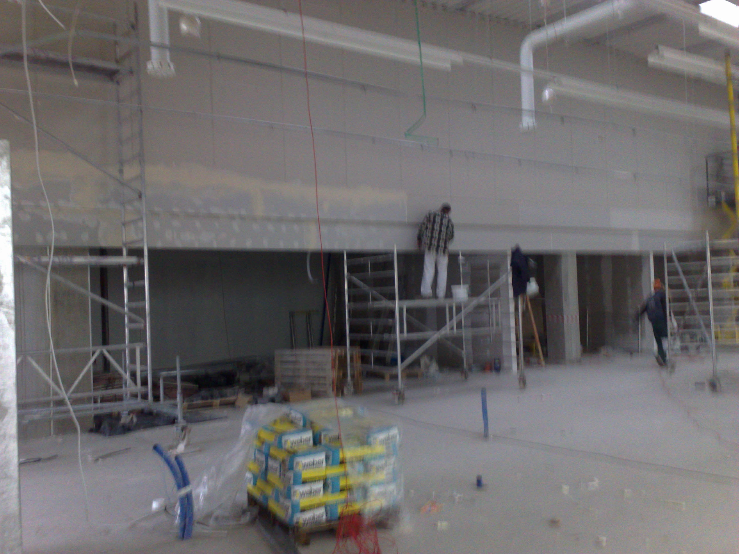 Montaż płyt gipsowo-kartonowych na dużej powierzchni ściany w hali, widoczne rusztowania, pracownicy, worki z zaprawą i instalacje pod sufitem.