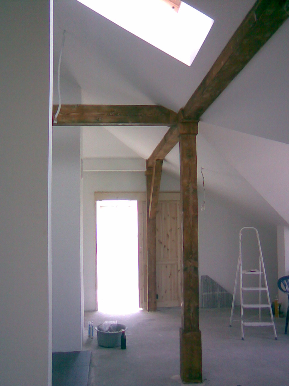 Poddasze z drewnianą konstrukcją nośną, widoczne elementy zabudowy z płyt G-K, drabina i wiadro z narzędziami, oświetlone oknem dachowym.