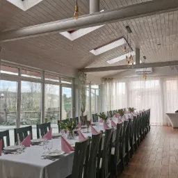 Uroczysta aranżacja długiego stołu w jasnej sali w Gorzycach, z widokiem na zieleń za oknem. Stół nakryty białym obrusem, z różowymi serwetkami, talerzami, kieliszkami i bukietami tulipanów...