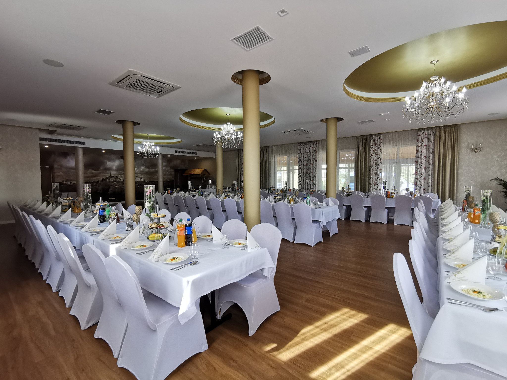 Sala bankietowa z długimi stołami, nakryta białymi obrusami i krzesłami w pokrowcach, przygotowana na przyjęcie, z widocznymi napojami, talerzami i dekoracjami.