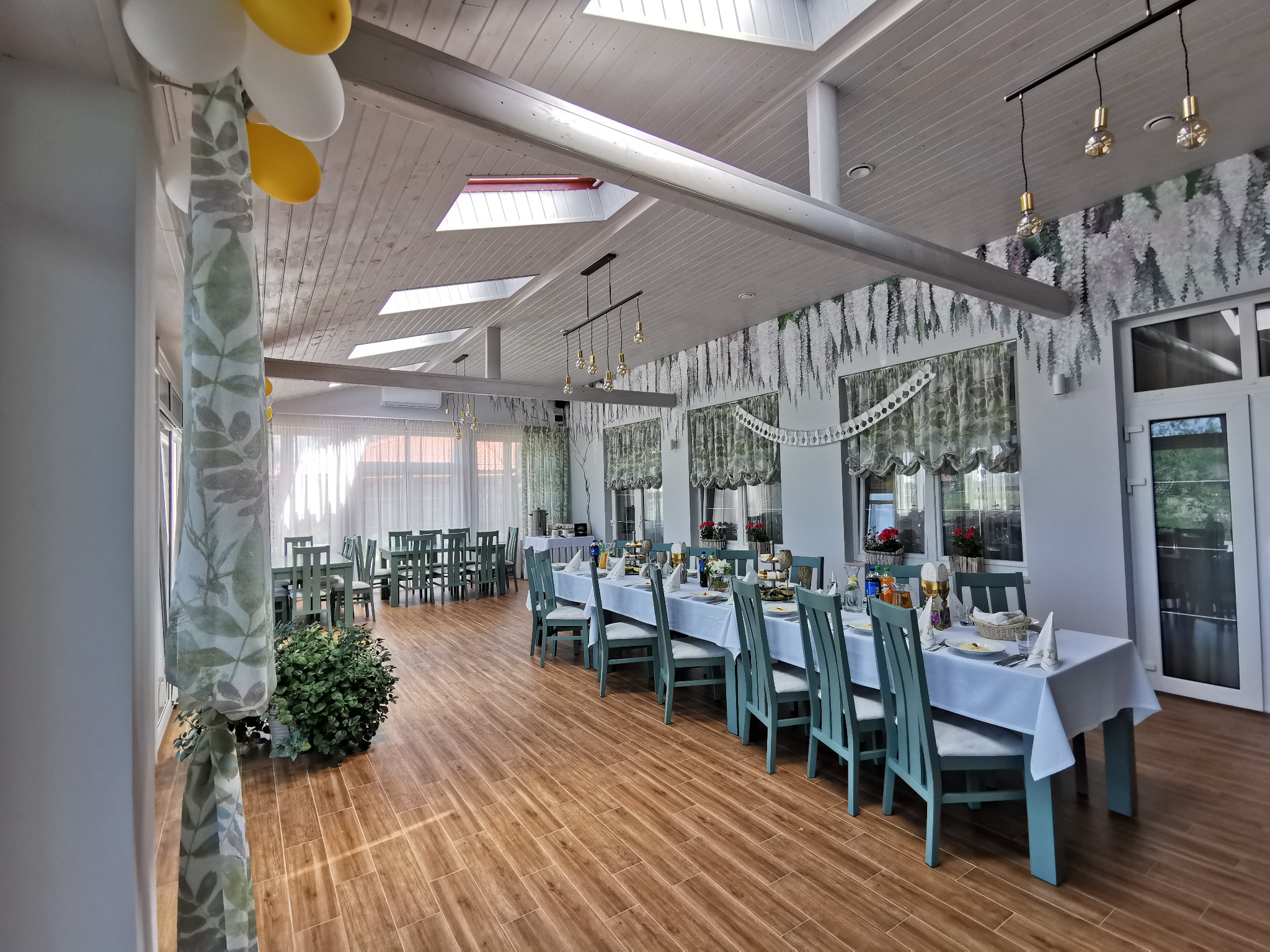 Sala bankietowa z nakrytymi stołami, krzesłami w kolorze morskim, dekoracjami kwiatowymi na ścianach i balonami pod sufitem, przygotowana na przyjęcie.