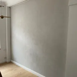 Ściana z efektem dekoracyjnym w odcieniach szarości, listwy przypodłogowe i sufitowe w bieli, parkiet w jasnym odcieniu drewna, widoczne gniazdka elektryczne przy podłodze.