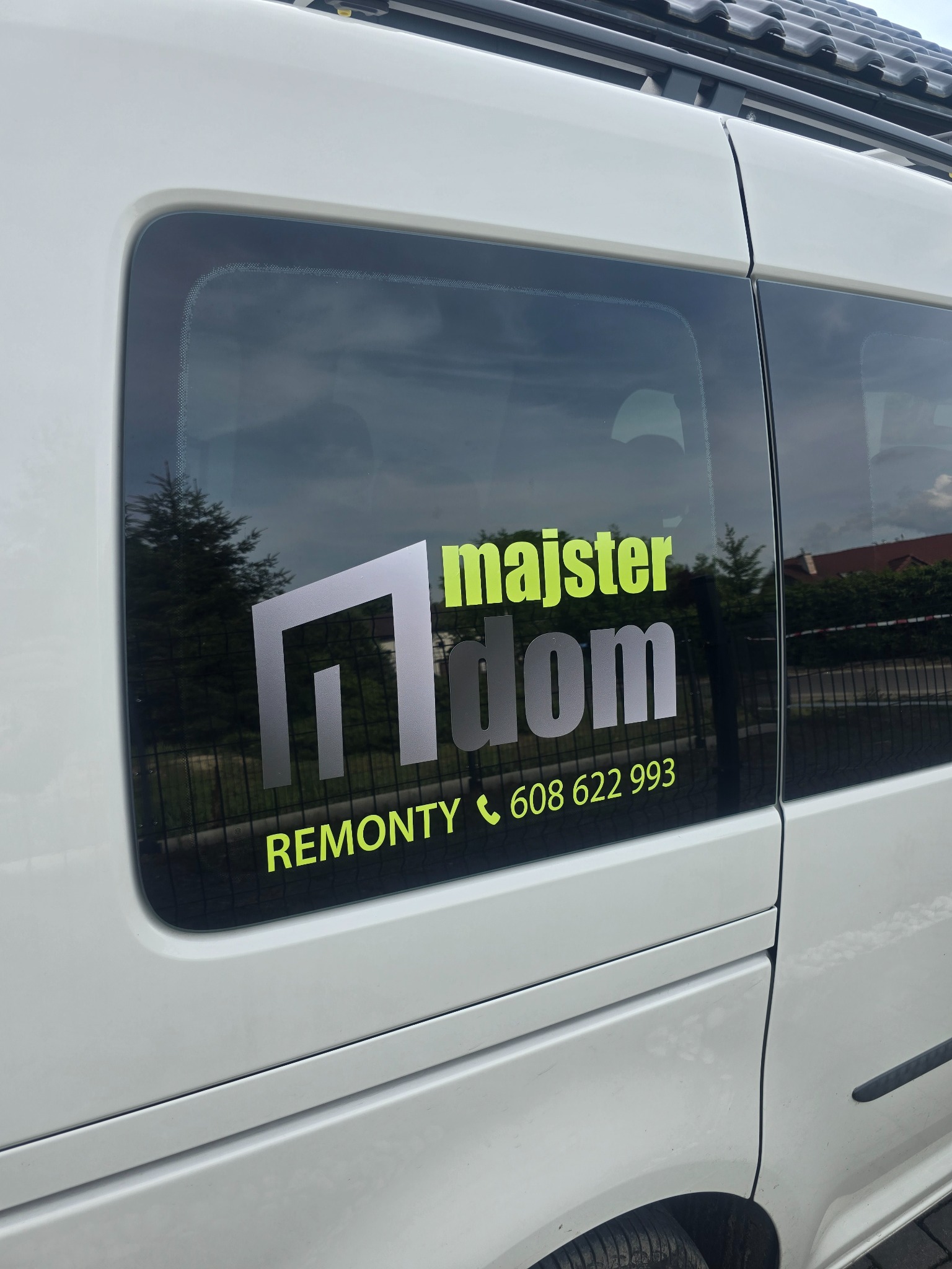 Biały samochód z naklejką 'majster dom' i numerem telefonu na szybie. Widoczne tło z zielenią i fragment dachu budynku. Reklama usług remontowych.