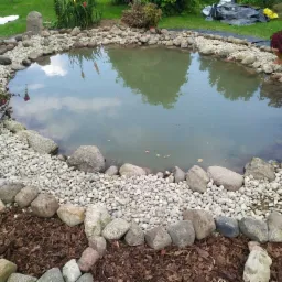 Oczko wodne z brzegiem wyłożonym kamieniami różnej wielkości i żwirem, otoczone korą drzewną, z odbiciem nieba w wodzie i dekoracyjnym koszykiem z kwiatami po lewej stronie.