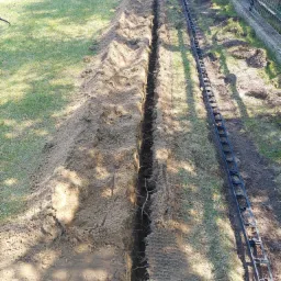 Wykopany wąski rów w trawniku, obok leży czarny element konstrukcyjny, prawdopodobnie do instalacji nawadniającej, widoczny fragment rury nawadniającej.