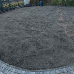 TB Kompleksowa Budowa Ogrodów - Przygotowany teren pod trawnik, wyrównana ziemia z widocznymi śladami po grabiach i rozsianymi nasionami trawy, obrzeże z kostki brukowej, pracownik w oddali.