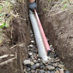TB Kompleksowa Budowa Ogrodów - Instalacja drenażu w Strzegocinie: Rów wykopany w ziemi, wypełniony kamieniami, w którym umieszczana jest perforowana rura drenażowa, poziomowana czerwoną poziomicą.