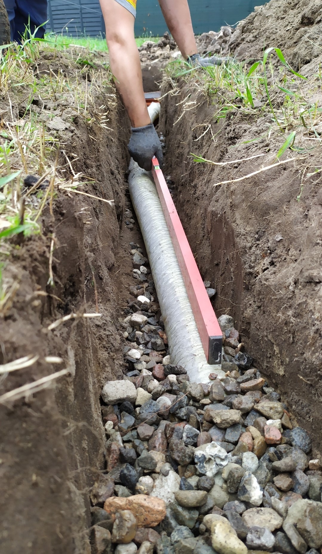 Instalacja drenażu w Strzegocinie: Rów wykopany w ziemi, wypełniony kamieniami, w którym umieszczana jest perforowana rura drenażowa, poziomowana czerwoną poziomicą.