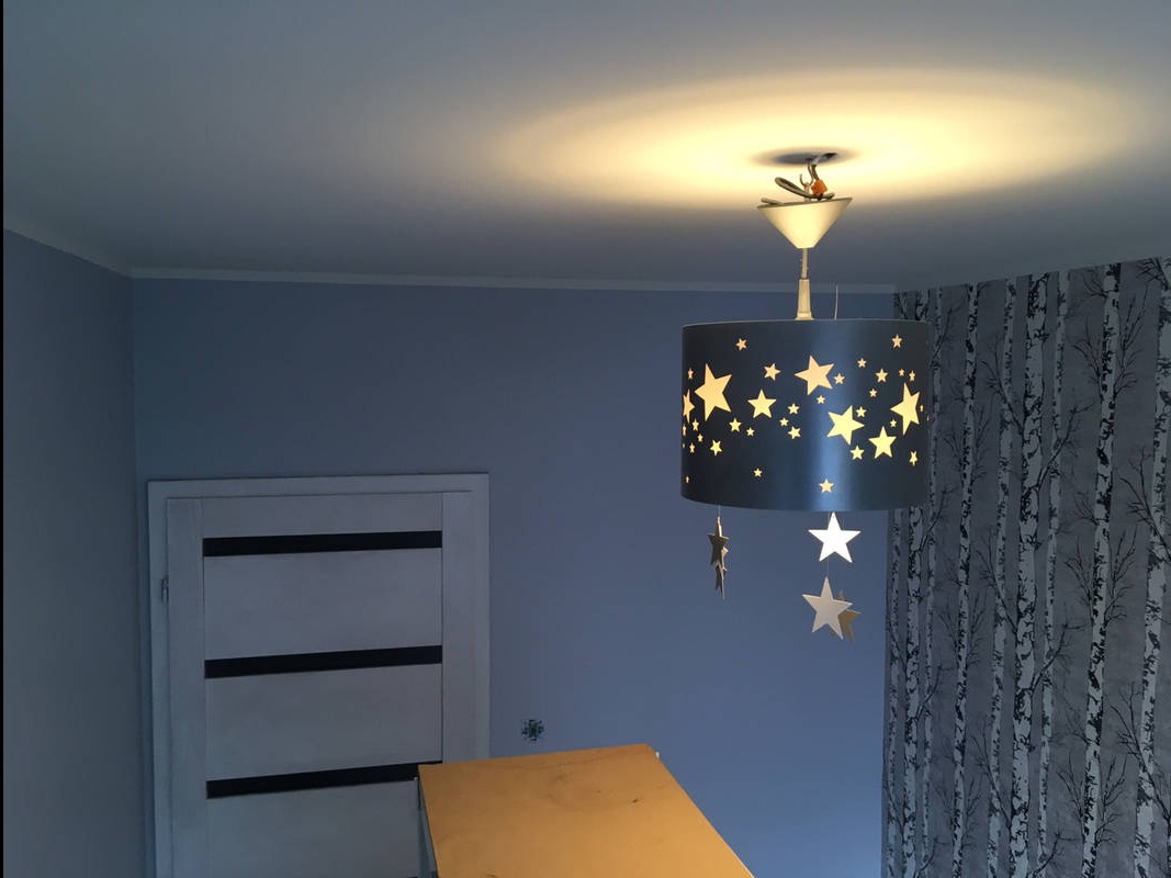 Niebieski pokój dziecięcy z tapetą w brzozy, lampą w gwiazdki i częściowo widocznym stołem roboczym. Świeżo wykończone wnętrze.