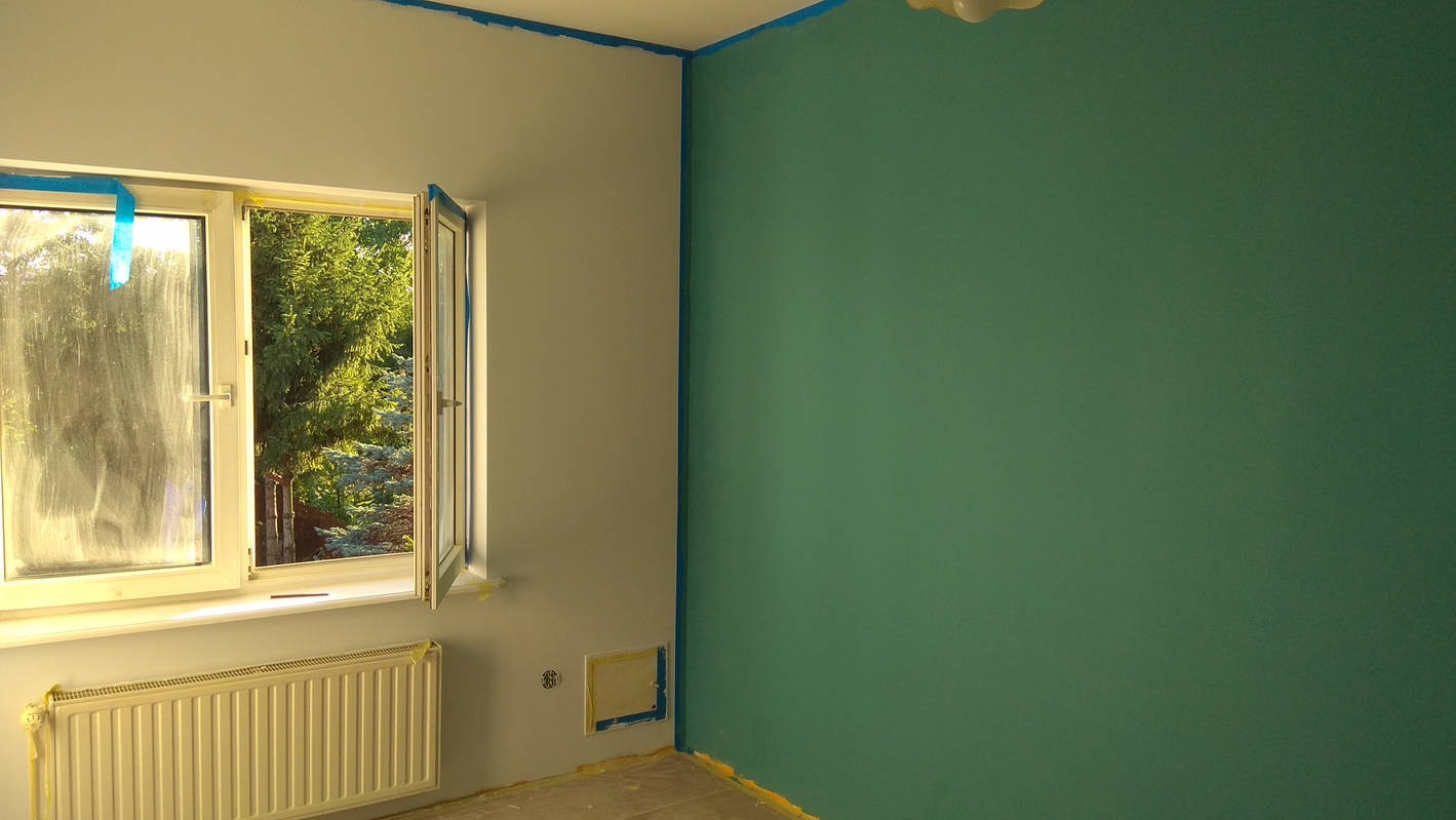 Świeżo pomalowane wnętrze z otwartym oknem, zabezpieczone taśmą malarską przy suficie i podłodze. Jedna ściana zielona, druga biała. Widok na zieleń za oknem.