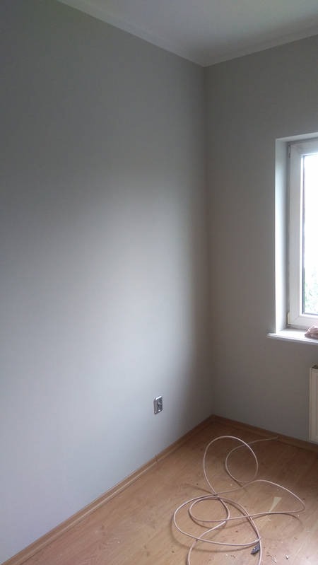 Jasnoszary pokój po odświeżeniu ścian, widoczne gniazdko elektryczne i fragment okna z parapetem. Na podłodze leżą zwinięte kable.