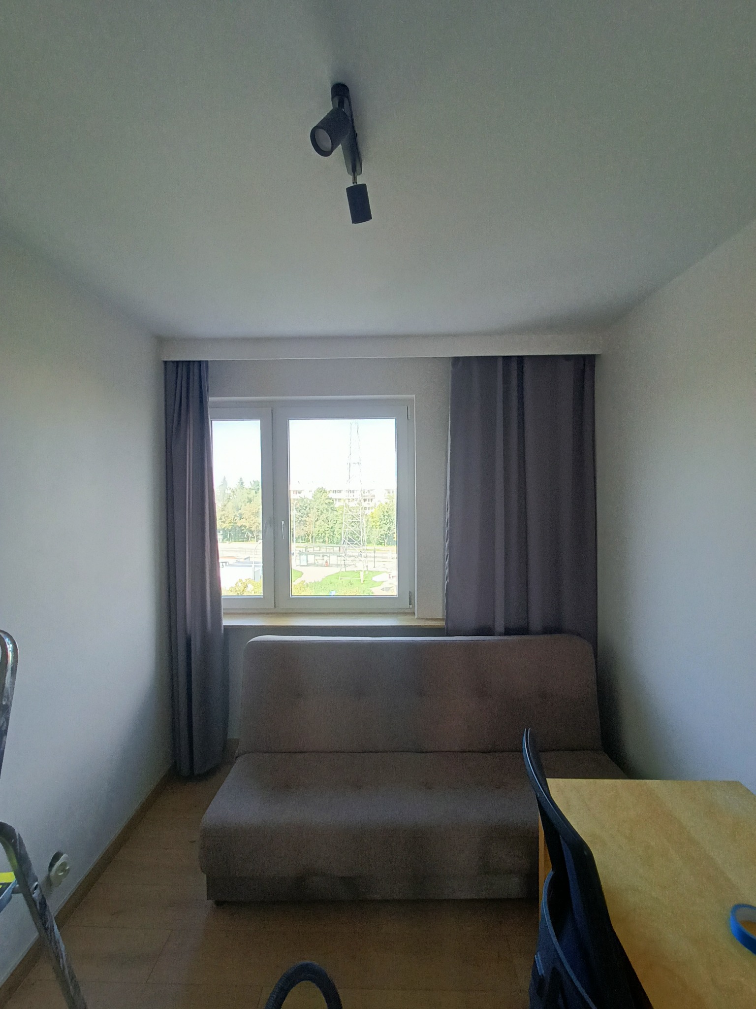 Wnętrze pokoju po odświeżeniu: jasne ściany, okno z szarymi zasłonami, sofa i fragment stołu z krzesłem. Widoczna drabina malarska w rogu.