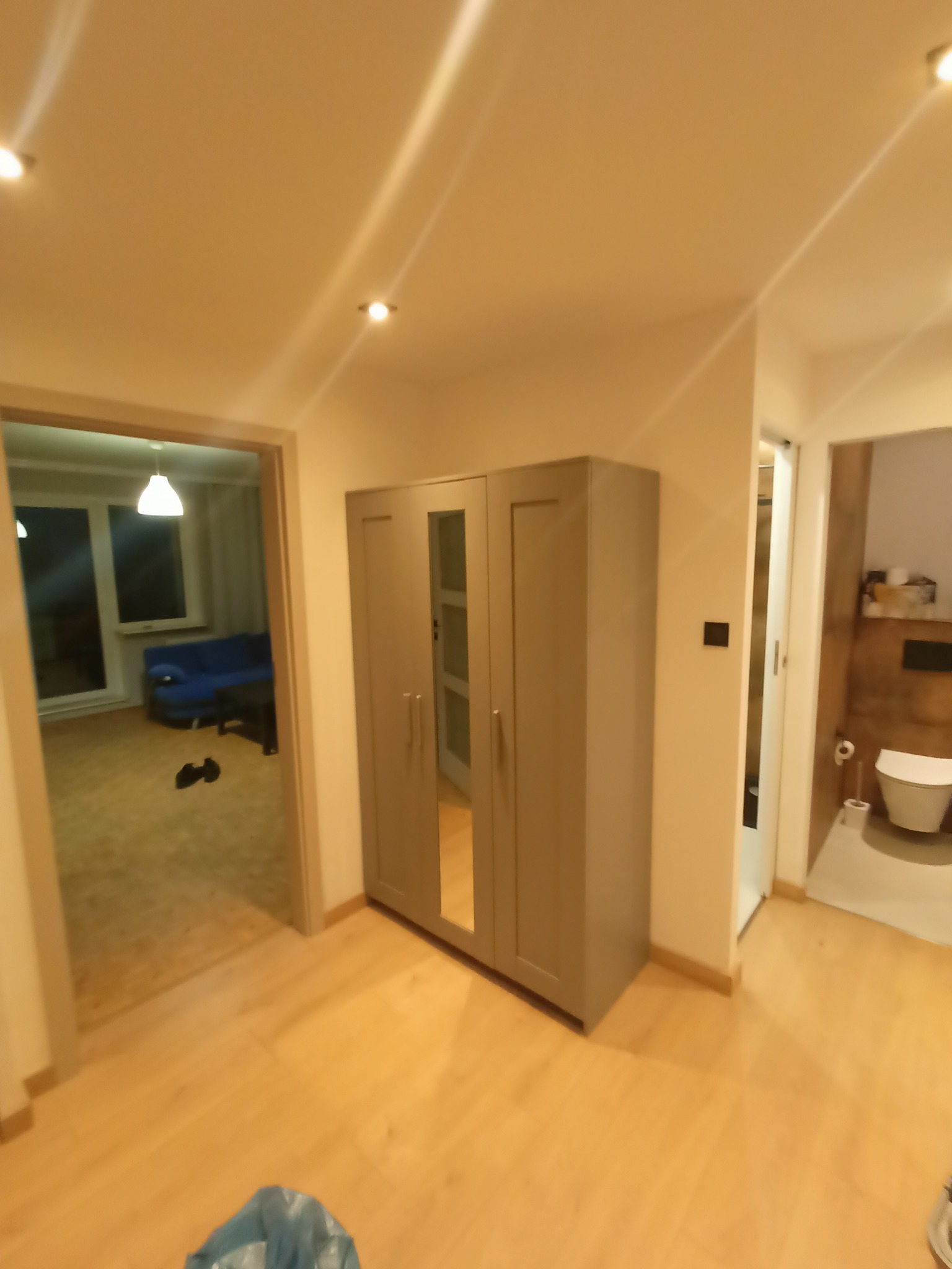 Wnętrze po malowaniu: świeżo pomalowane ściany w odcieniach beżu, widoczna szafa z lustrem, fragment łazienki i pokoju z niebieską sofą w tle. Podłoga wyłożona panelami.