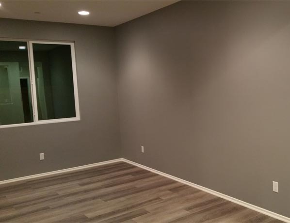 Pomieszczenie po remoncie: szare ściany, panele podłogowe imitujące drewno, białe listwy przypodłogowe i okno z widokiem na inne pomieszczenie.