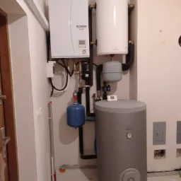 Zestaw grzewczy w kotłowni: Pompa ciepła Mitsubishi Electric Ecodan, biały zasobnik C.W.U. Galmet, szary bufor ciepła i niebieskie naczynie przeponowe, widoczne rury instalacyjne i osprzęt, ściany...