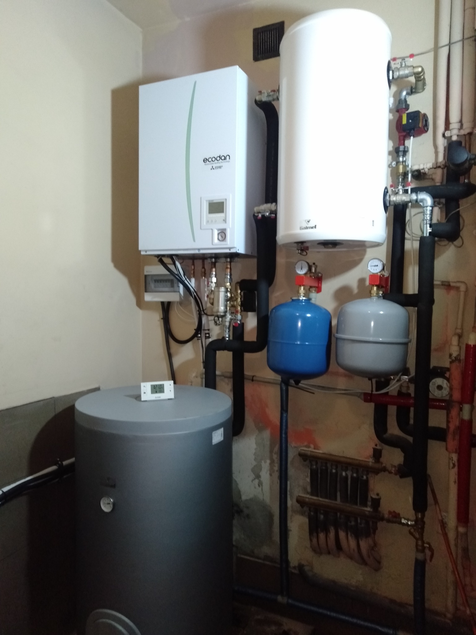 Kotłownia po modernizacji: Pompa ciepła Mitsubishi Electric Zubadan 8,0kW zamontowana na ścianie, obok biały zasobnik C.W.U. Salmet 200l, szary bufor 100l na podłodze i termostat pokojowy...