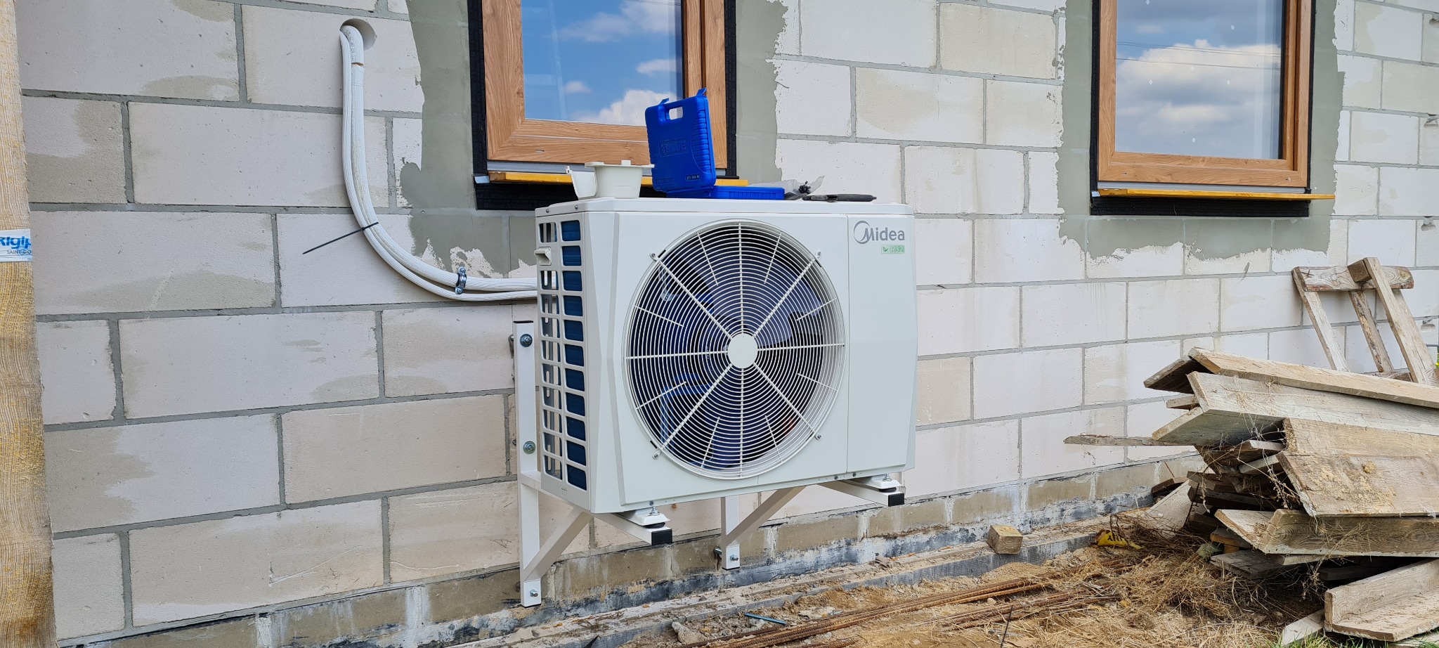 Biała jednostka zewnętrzna Midea M-Thermal II zamontowana na ścianie z białego bloczka, widoczne okna z drewnianą ramą, narzędzia na górze urządzenia i stos desek obok.