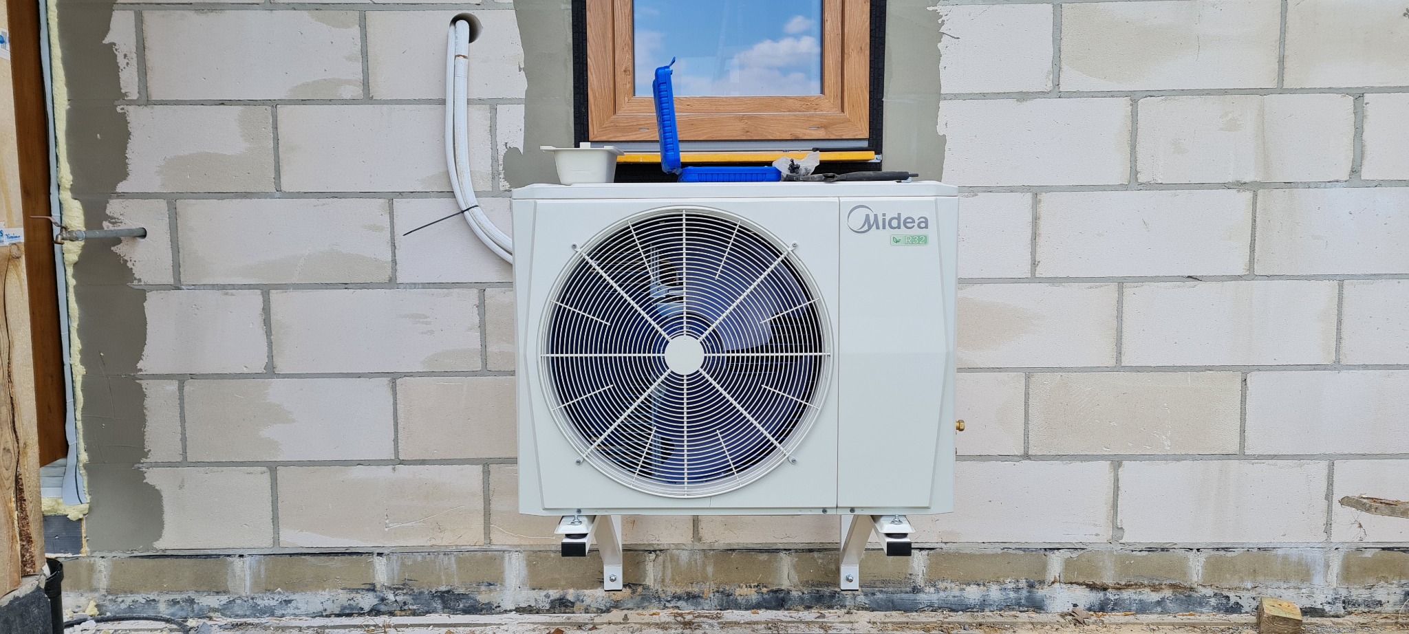 Biała jednostka zewnętrzna Midea z widocznym logo R32 zamontowana na wspornikach przy ścianie z białych bloczków, obok okna z drewnianą ramą, widoczne narzędzia montażowe.