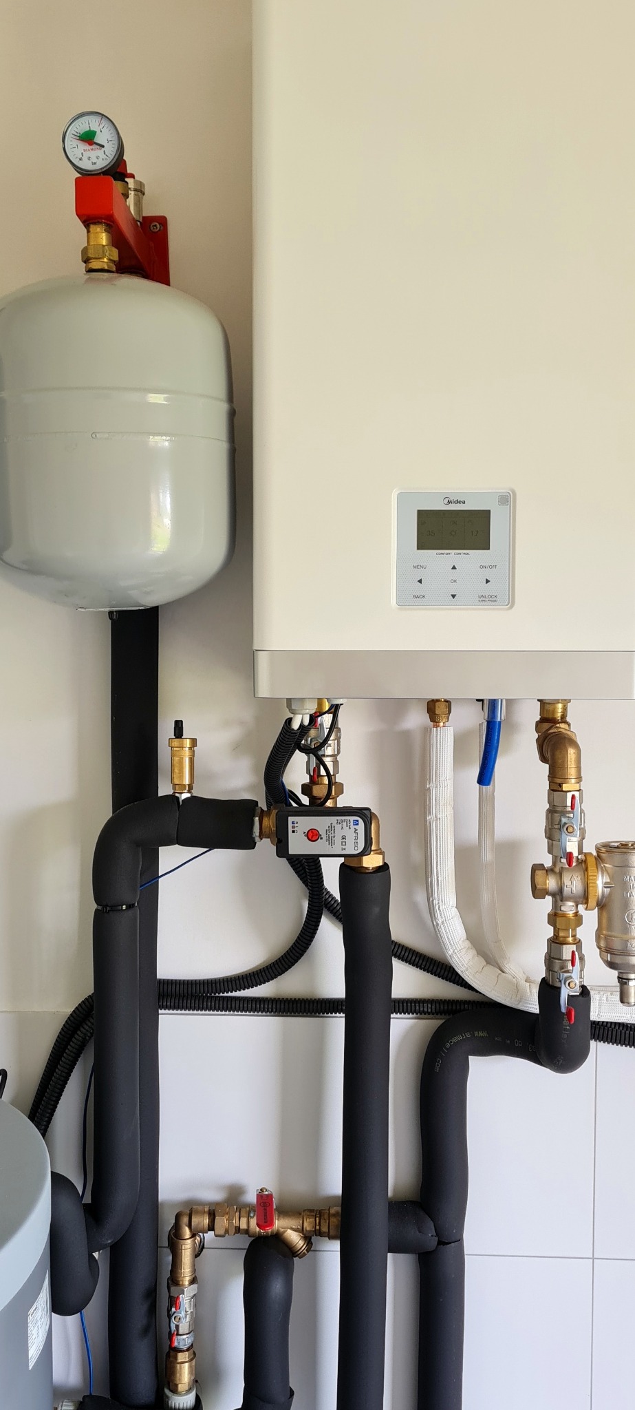 Zasobnik C.W.U. z manometrem na czerwonym wsporniku oraz moduł sterujący Midea z wyświetlaczem, połączone rurami w izolacji, widoczne elementy instalacji z zaworami i czujnikiem Afriso.