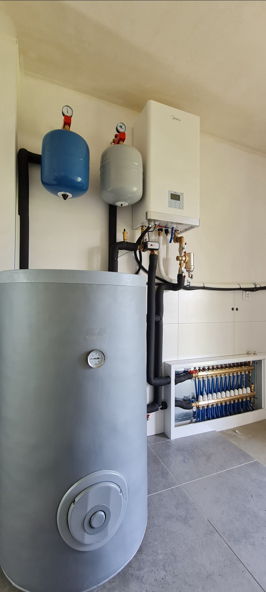 Kompletna instalacja pompy ciepła Midea M-Thermal II z zasobnikiem C.W.U. Galmet, widoczny rozdzielacz podłogowy z niebieskimi rurami, dwa naczynia wzbiorcze - niebieskie i szare, całość w jasnym...