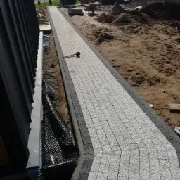 Długa ścieżka z jasnoszarej kostki granitowej ułożona przy budynku, widoczne warstwy drenażowe przy fundamentach, w tle ziemia, taczka i drewniana sauna.